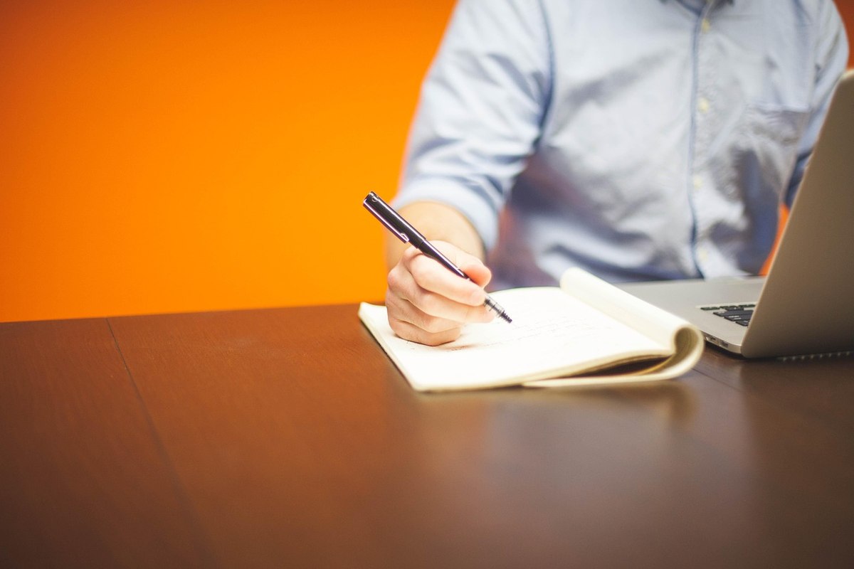 Personne assise à un bureau écrit dans un carnet, ordinateur portable à proximité, fond orange