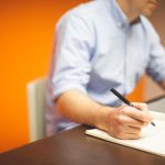 Homme en chemise bleue écrivant sur un bloc-notes, ordinateur portable ouvert sur son bureau, fond orange