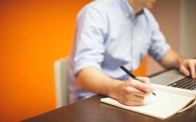 Homme en chemise bleue écrivant sur un bloc-notes, ordinateur portable ouvert sur son bureau, fond orange