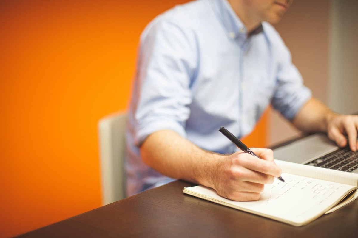 Homme en chemise bleue écrivant sur un bloc-notes, ordinateur portable ouvert sur son bureau, fond orange